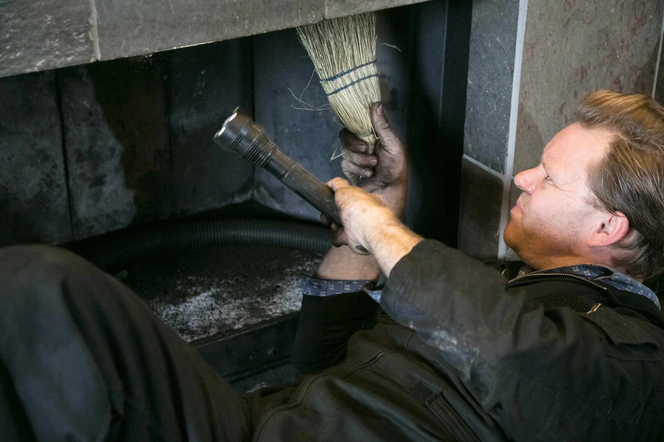 A man cleaning a fireplace holding a brush and flashlight.